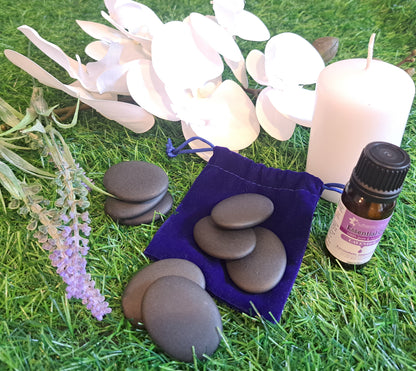 Eye-level product shot of lavender essential oil, a purple velvet bag, massage stones, a candle, and lavender sprigs on artificial turf.