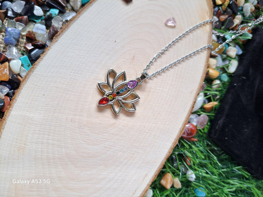 A silver lotus flower chakra pendant necklace on a wooden surface surrounded by colorful stones and artificial grass.