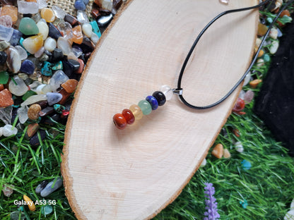 A chakra crystal necklace rests on a wooden slice, surrounded by loose gemstones and artificial grass.